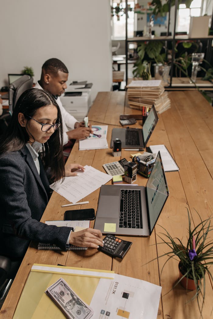 Two diverse accountants working at a shared office table using laptops and calculators.